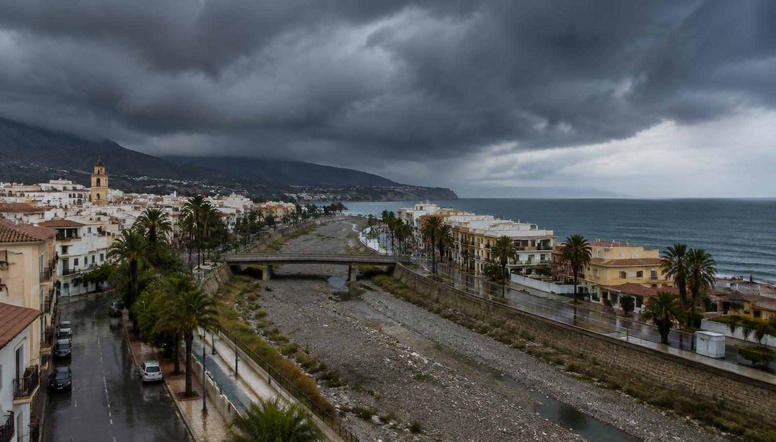 Dunkle Regenwolken über einer spanischen Mittelmeerstadt mit trockenem Flussbett (KI-generiertes Symbolbild)