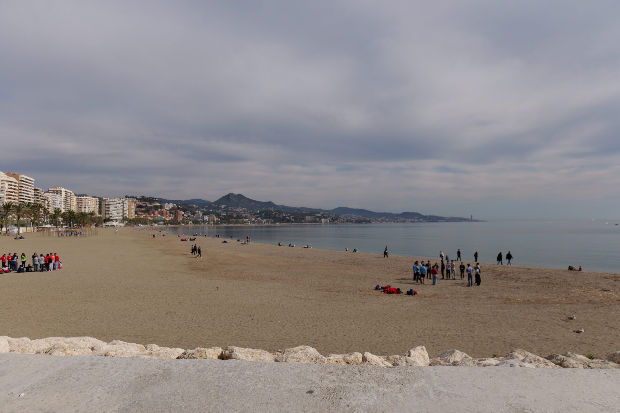 Breiter Sandstrand von Málaga mit Blick auf die Stadt, das ruhige Meer und Spaziergänger unter leicht bewölktem Himmel.